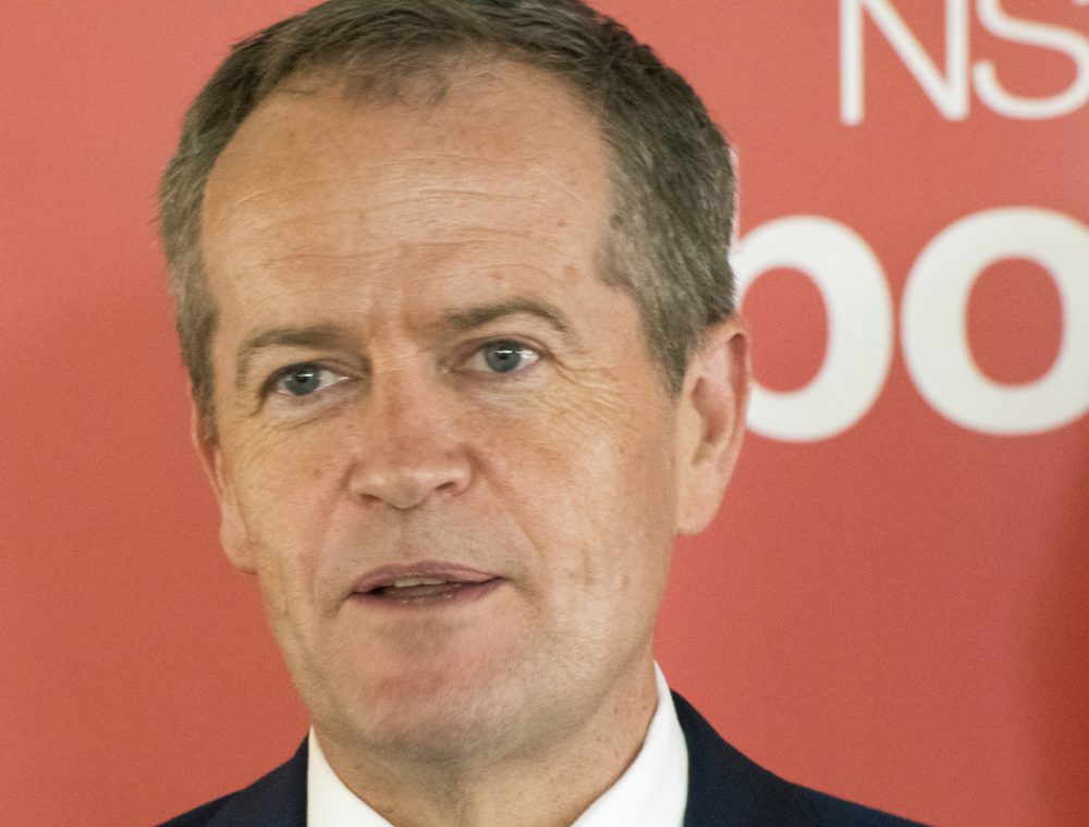 NDIS Minister Bill Shorten, a white middle aged man, in front of a red background with white text on it.