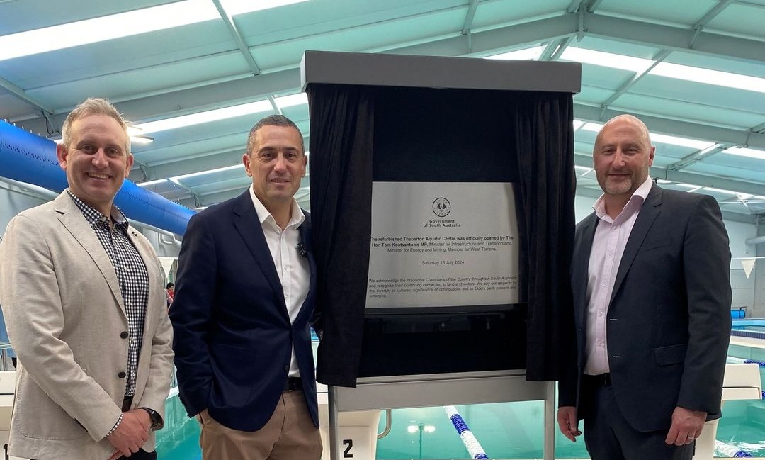 Three men stand before a plaque to open the new aquatic centre. They are dressed in suit jackets and smiling. There is a swimming pool behind them.