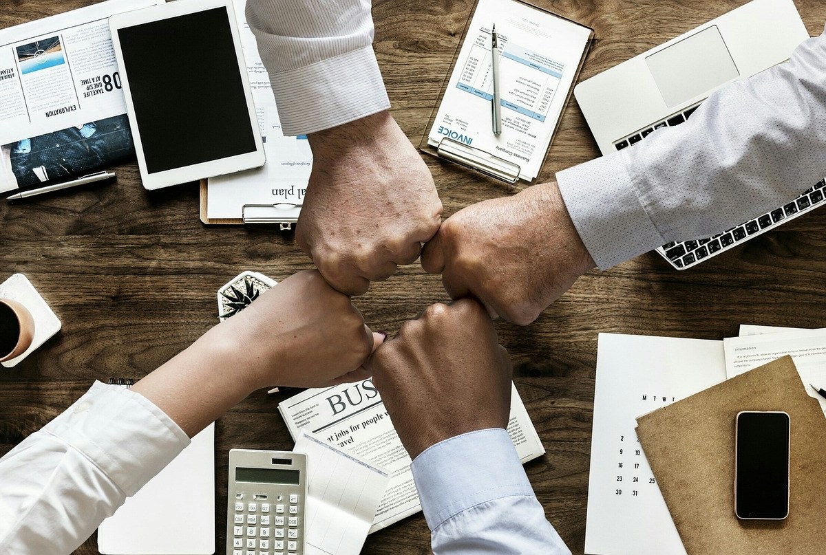 Four arms reach across a table to bump fists. They are wearing business shirts and the table is covered in business materials and papers.