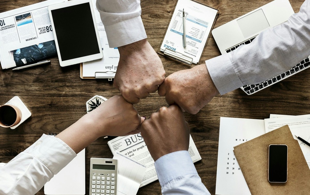 Four arms reach across a table to bump fists. They are wearing business shirts and the table is covered in business materials and papers.