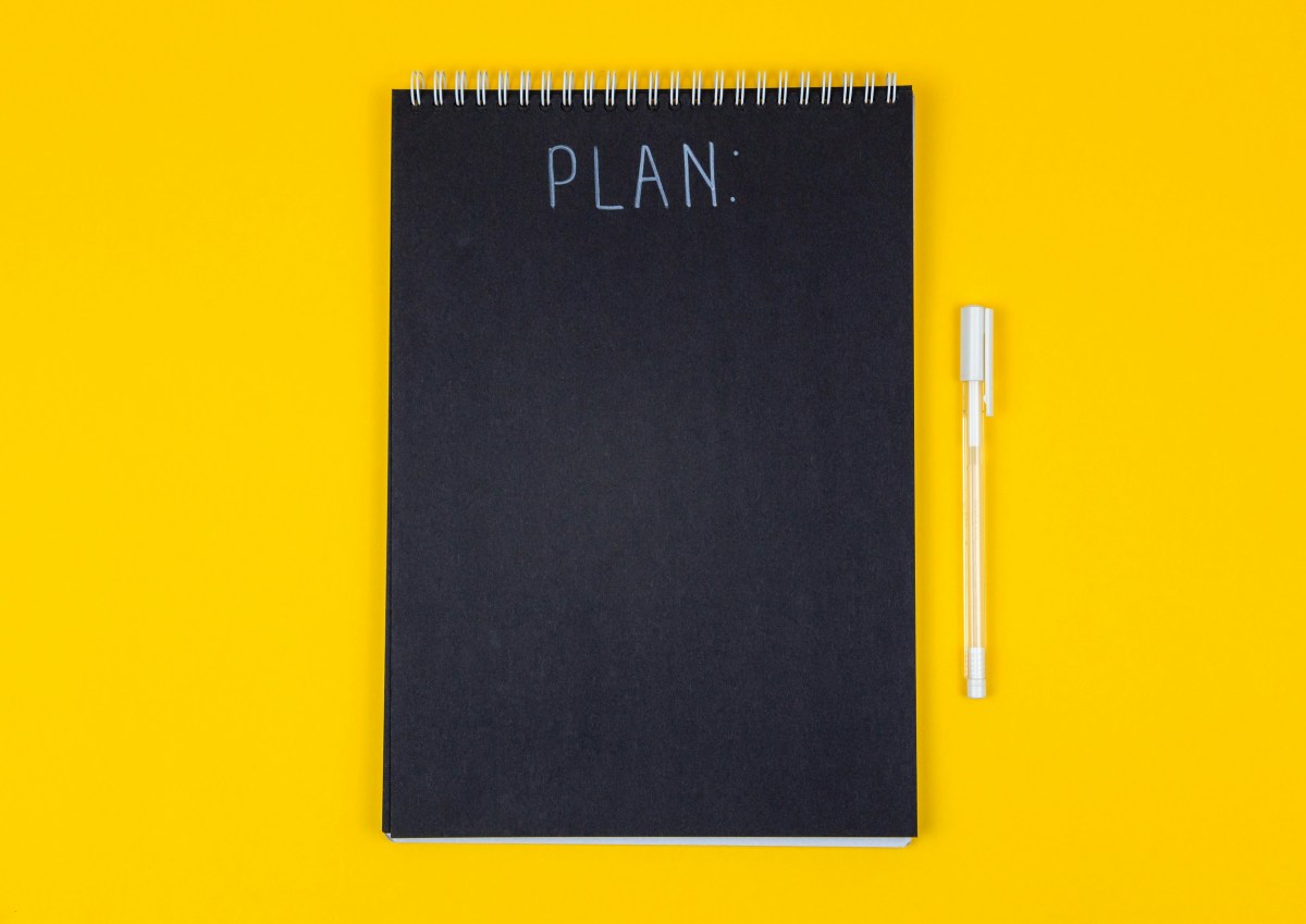 A black notepad on a yellow background with a white pen. The word 'plan' is written in white on the notepad at the top.