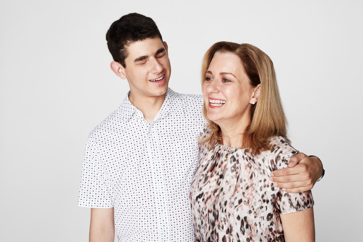 A teenage boy with dark hair wearing a pale shirt, with his arm around his mum, a middle aged woman with blonder hair and a patterned shirt.