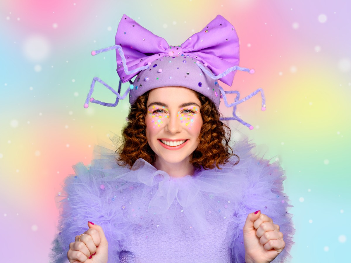 A young woman with brown shoulder length hair wearing a purple fluffy costume, with a large purple bow in her hair. She is smiling brightly. She is an actor in Fancy Long Legs.