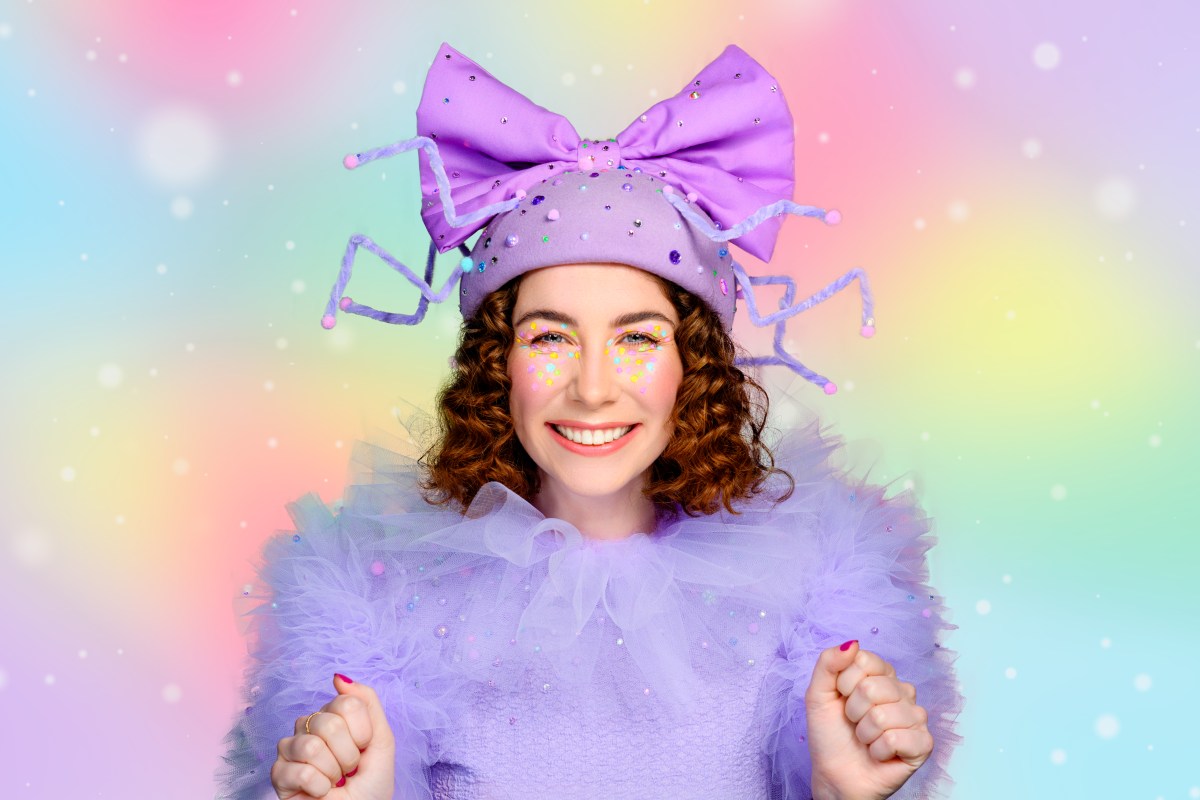 A young woman with brown shoulder length hair wearing a purple fluffy costume, with a large purple bow in her hair. She is smiling brightly. She is an actor in Fancy Long Legs.