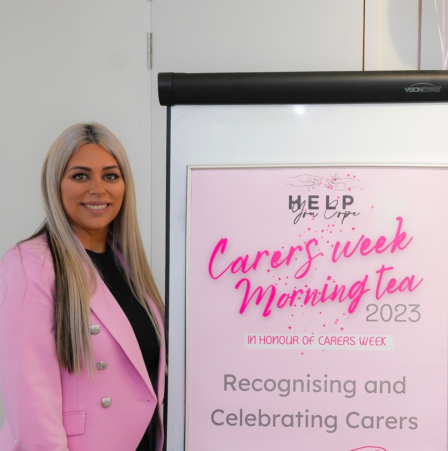 Joline George, founder of Help You Cope. She has long blonde hair and is wearing a pink blazer, and is standing next to a banner about a morning tea.