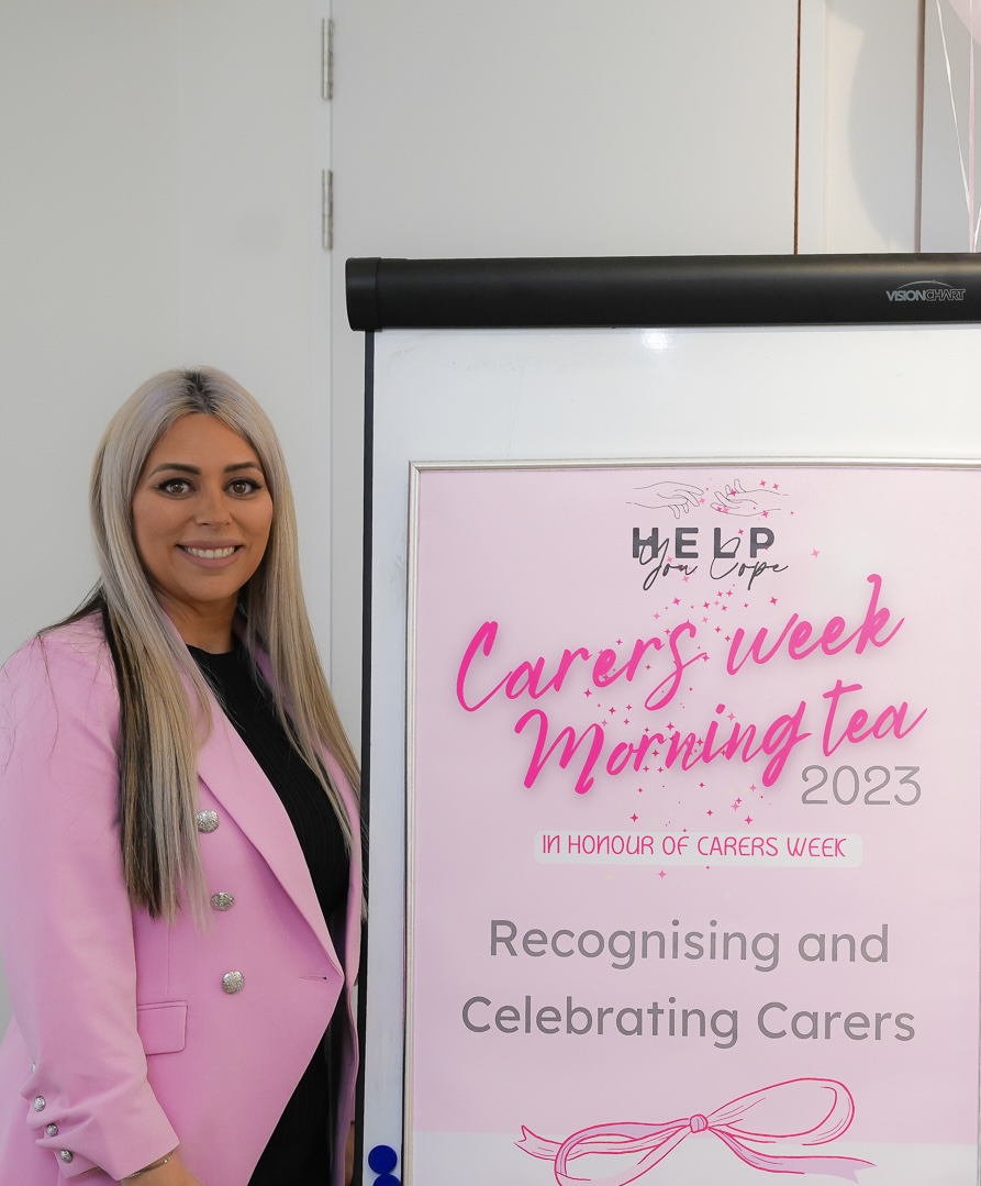 Joline George, founder of Help You Cope. She has long blonde hair and is wearing a pink blazer, and is standing next to a banner about a morning tea.