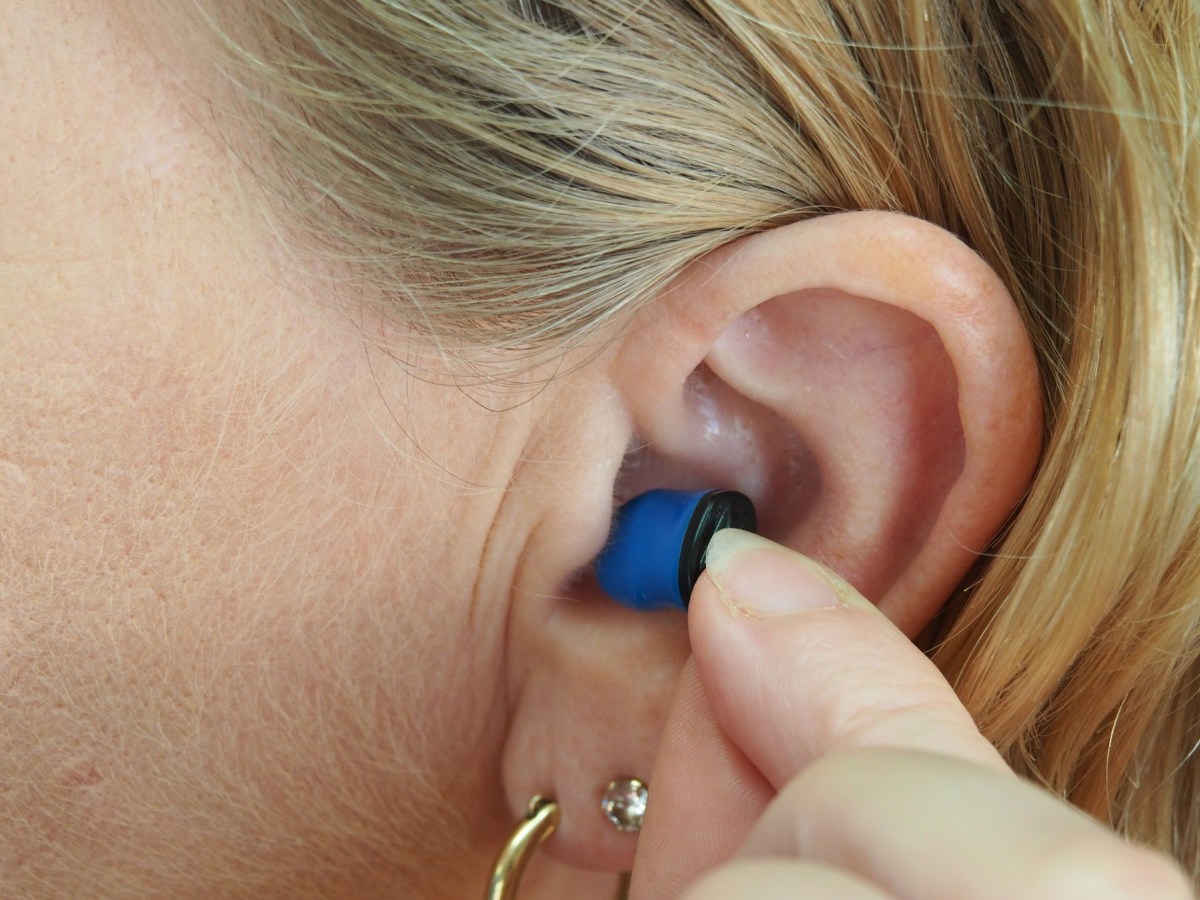 We see a close up shot of a woman putting a tiny hearing aid in her ear. It is blue and she has blonde hair tucked behind her ear.