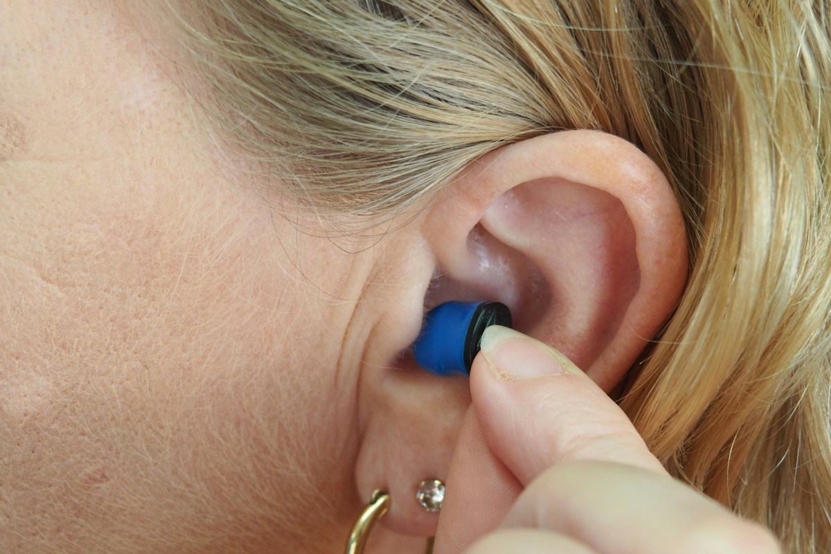 We see a close up shot of a woman putting a tiny hearing aid in her ear. It is blue and she has blonde hair tucked behind her ear.