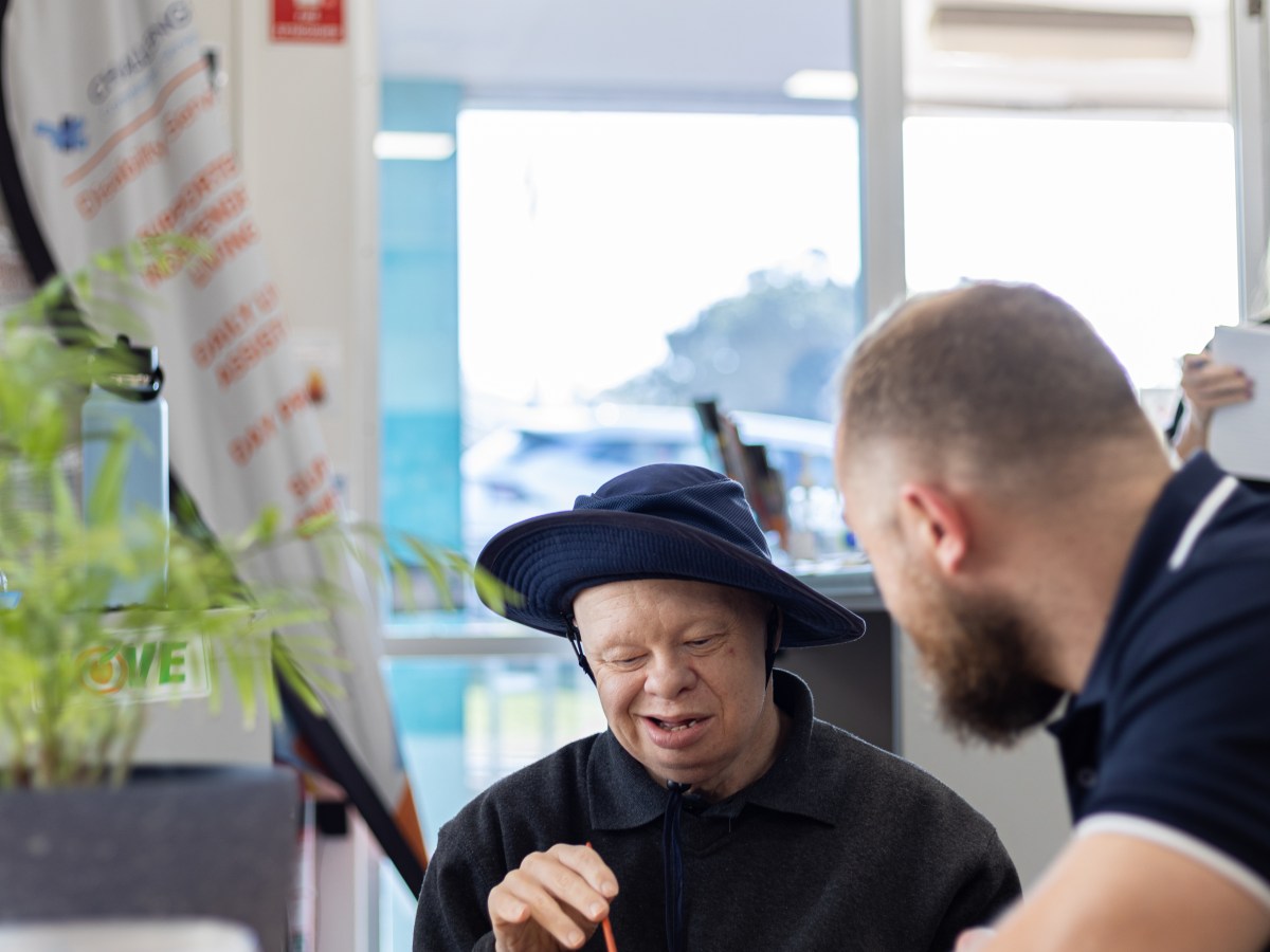 Two people sit at a table. One, a person with a visible disability, is painting and facing the camera. The other, an older man with a beard, is facing the first person.