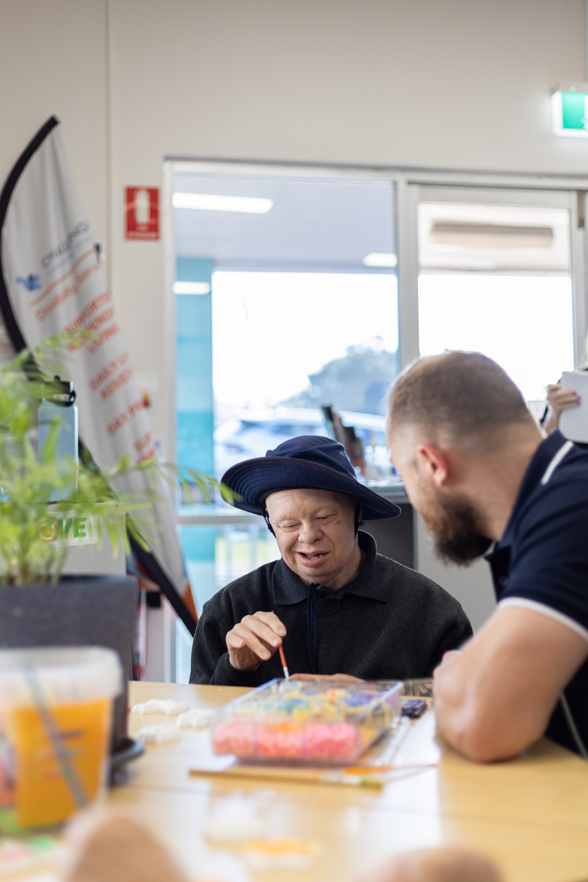 Two people sit at a table. One, a person with a visible disability, is painting and facing the camera. The other, an older man with a beard, is facing the first person.