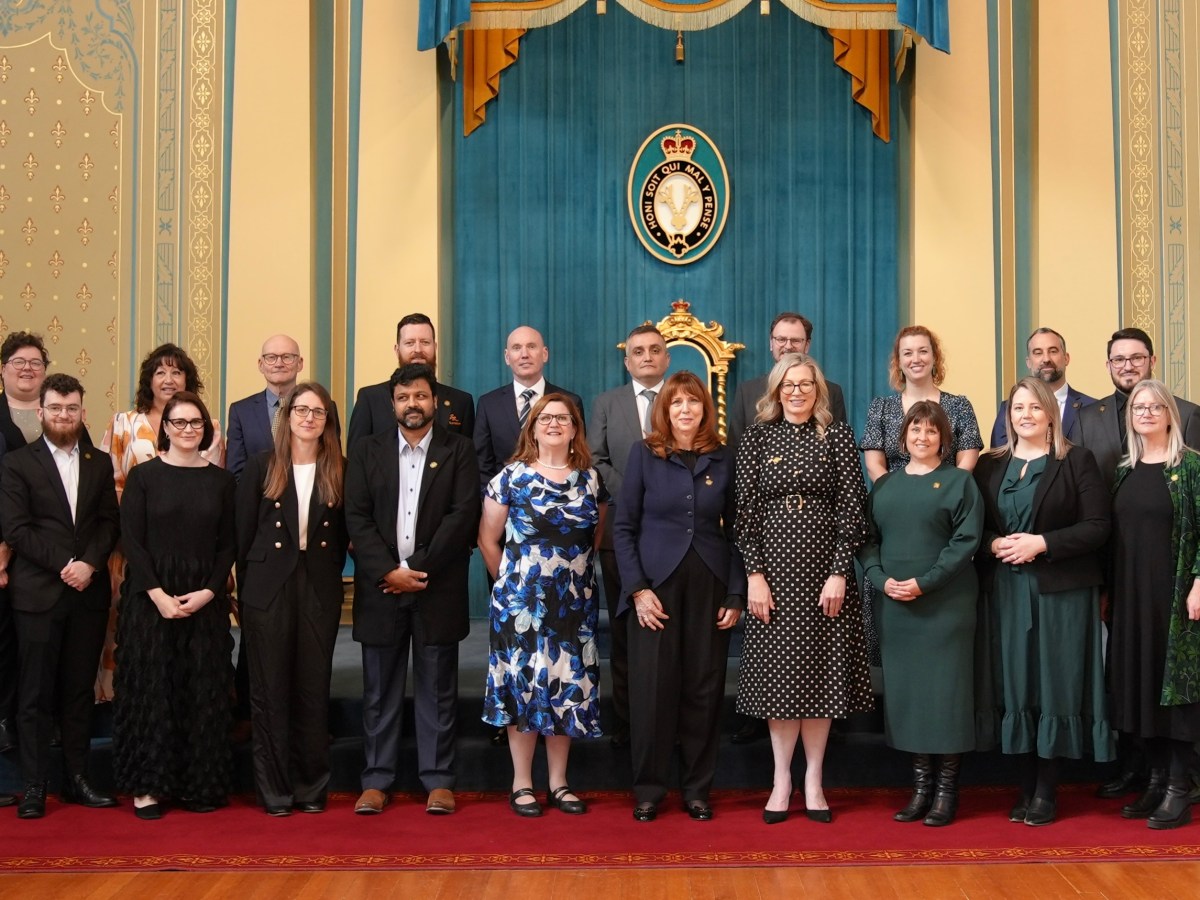 A group of people, all in suits and professional dress, stand for an official portrait on a red floor in front of a teal and yellow background.