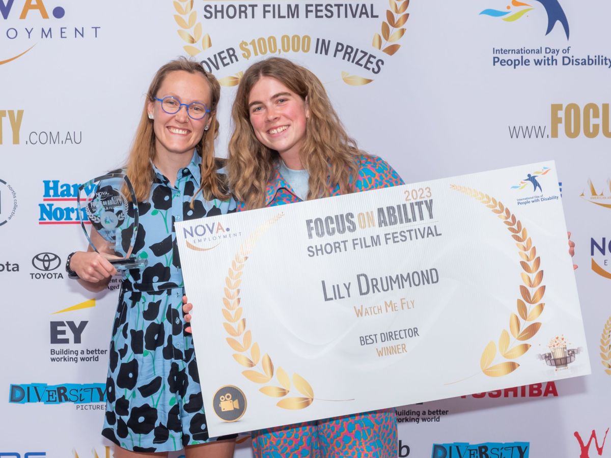 Two women in front of a wall with the film festival logo on it, one of them holding a large sign about the winner.