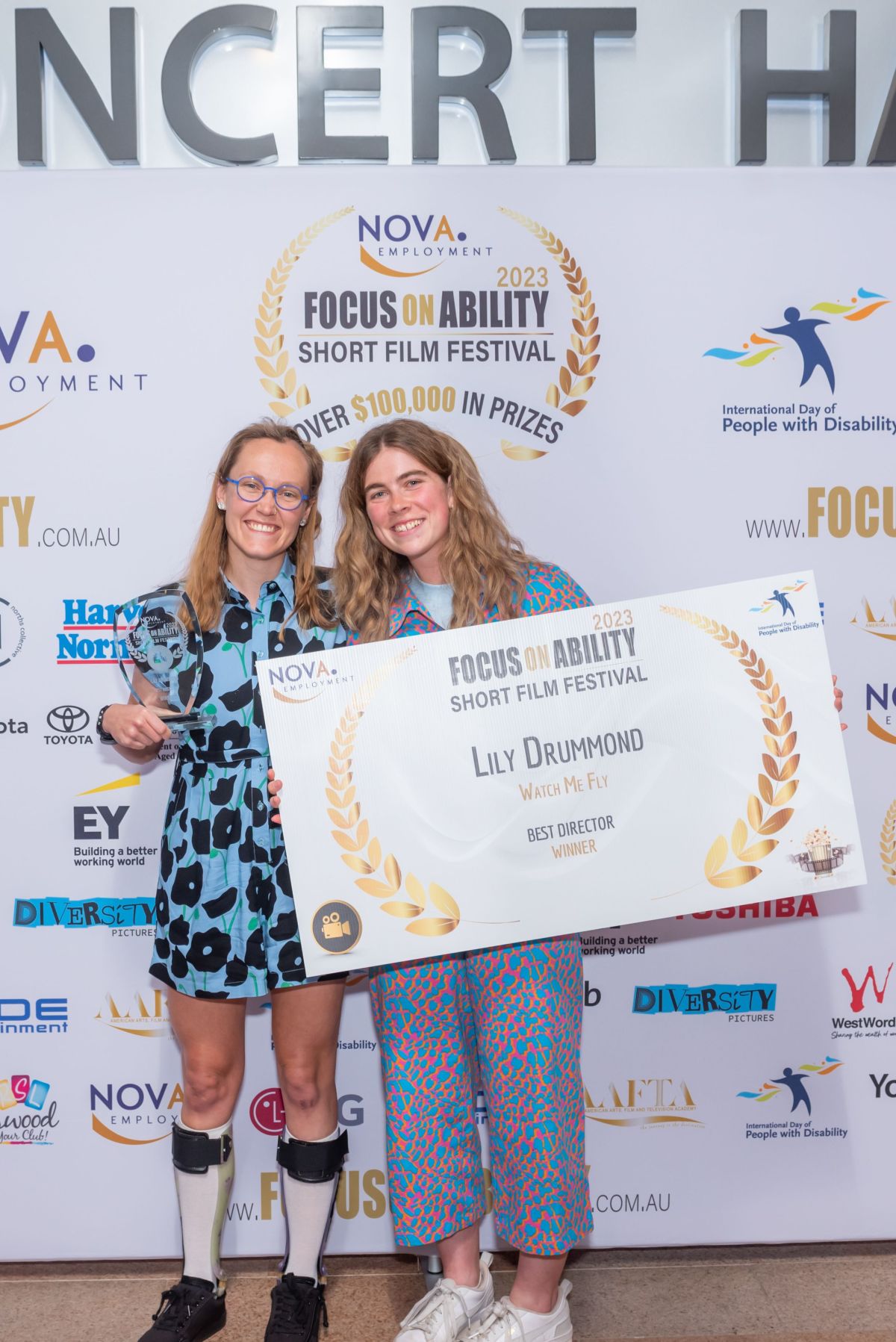 Two women in front of a wall with the film festival logo on it, one of them holding a large sign about the winner.