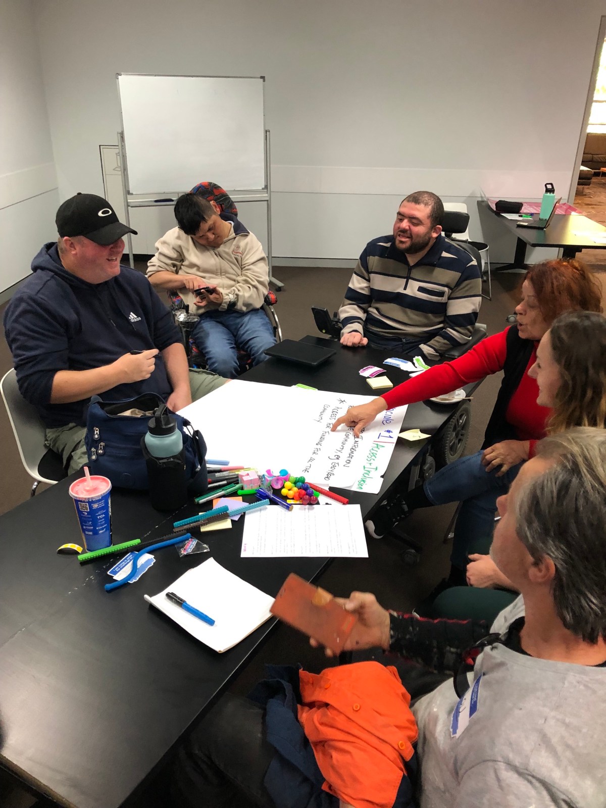 A group of people, some with a physical disability, sit in a room around a table with documents on it. They are having a discussion.