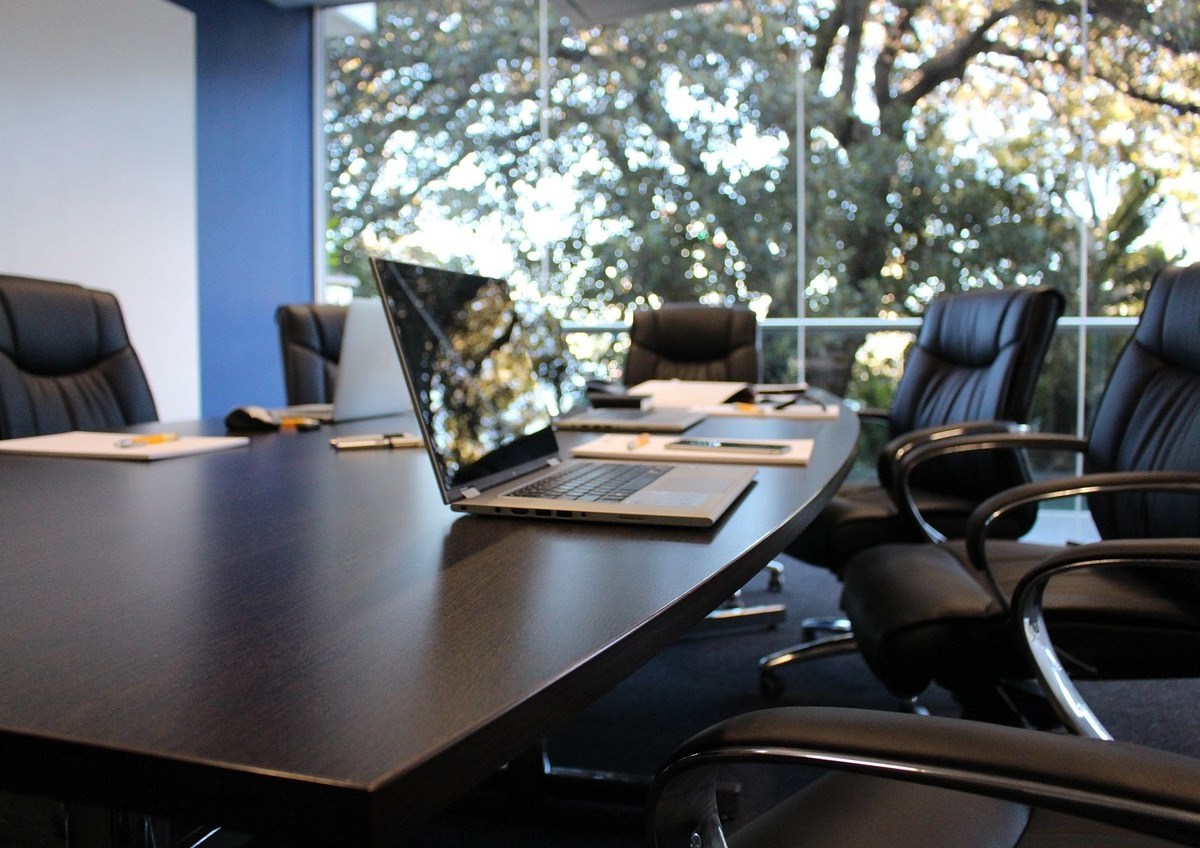 A boardroom featuring black leather chairs around a polished table, with a laptop on it. There are trees outside the window.