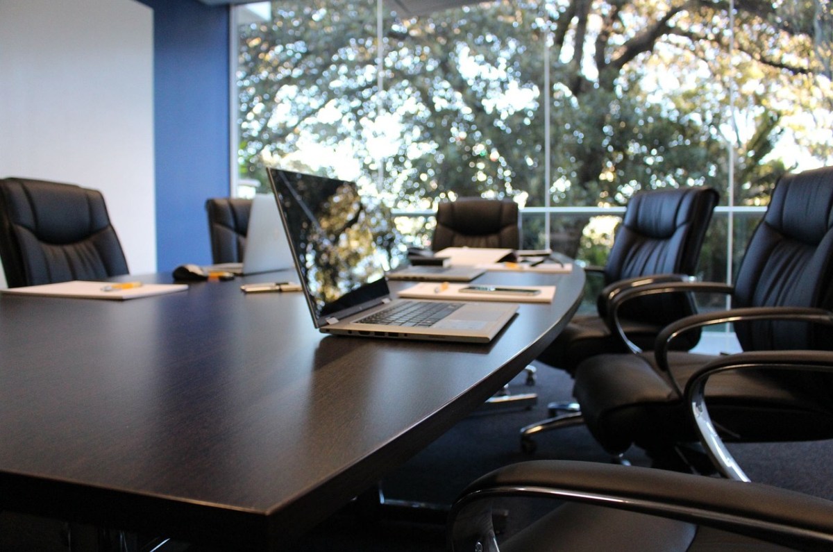 A boardroom featuring black leather chairs around a polished table, with a laptop on it. There are trees outside the window.