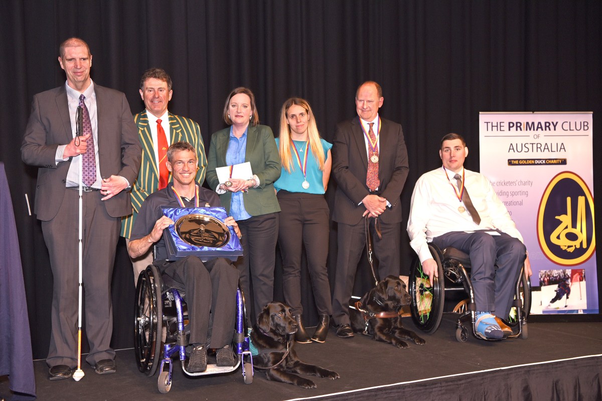 A group of people at a previous Primary Club dinner. They are on a stage and several people have a visible disability, including two people in a wheelchair and one who uses a white cane.