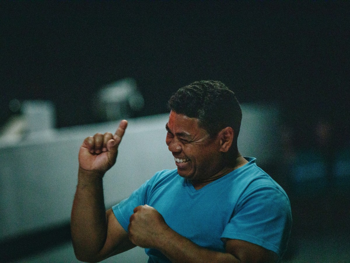 A dark-skinned man in a blue tshirt signs and smiles.