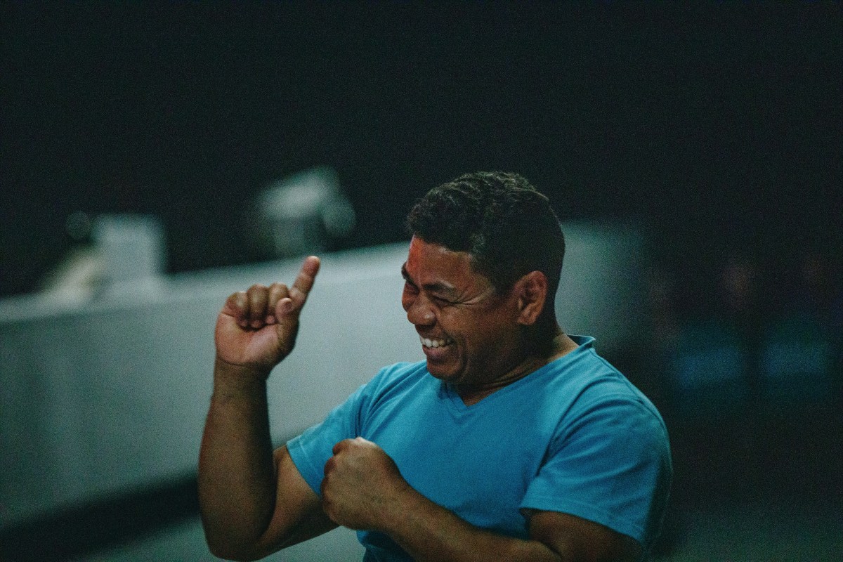 A dark-skinned man in a blue tshirt signs and smiles.