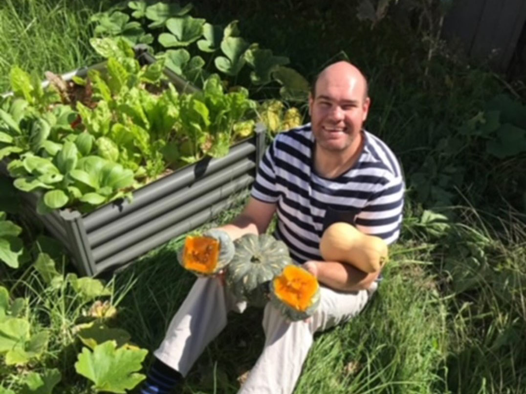 Andrew Radford, a beaming young man in a striped shirt, sits next to a garden bed holding veggies and flowers.