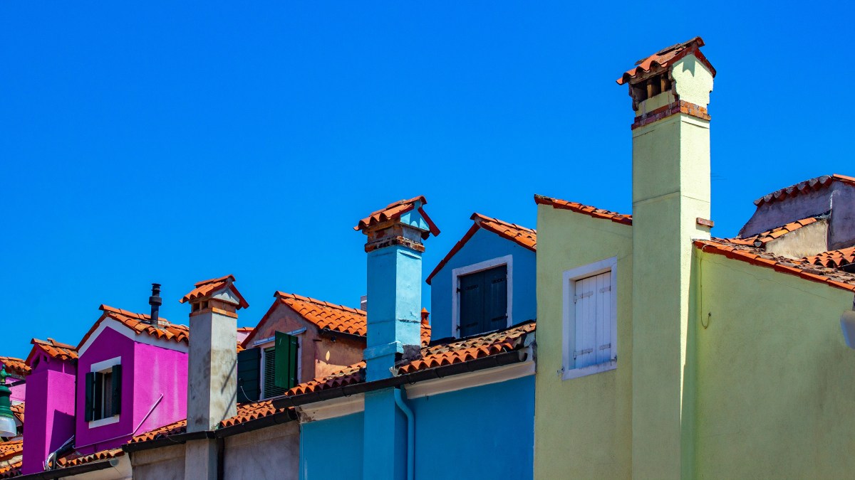 A row of connected houses in bright colours like pink, blue and green. We can only see the top of each house. The sky behind them is bright blue.