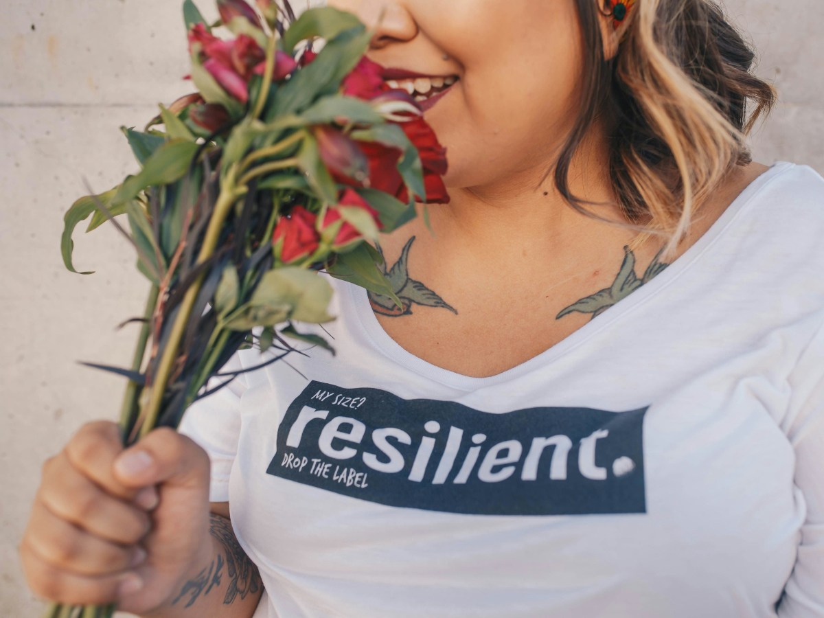 A lady wears a white shirt that says 'resilience' in big letters. She is sniffing a bunch of flowers.