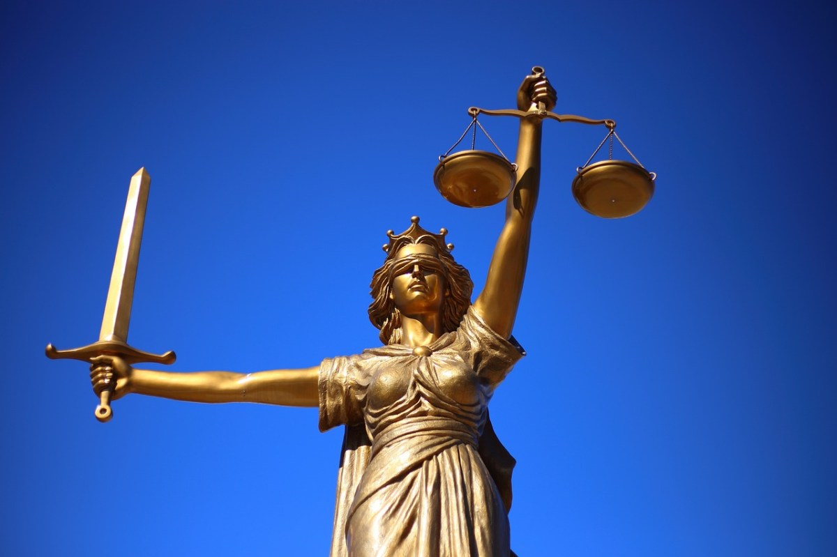 A photo of a gold statute of justice, a blindfolded woman holding a sword and the scales of justice, against a bright blue sky.