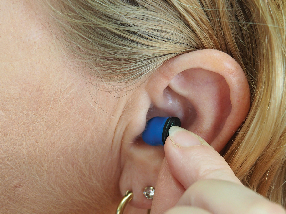 We can see a woman's ear. She is pointing to and touching a blue hearing aid in her ear.