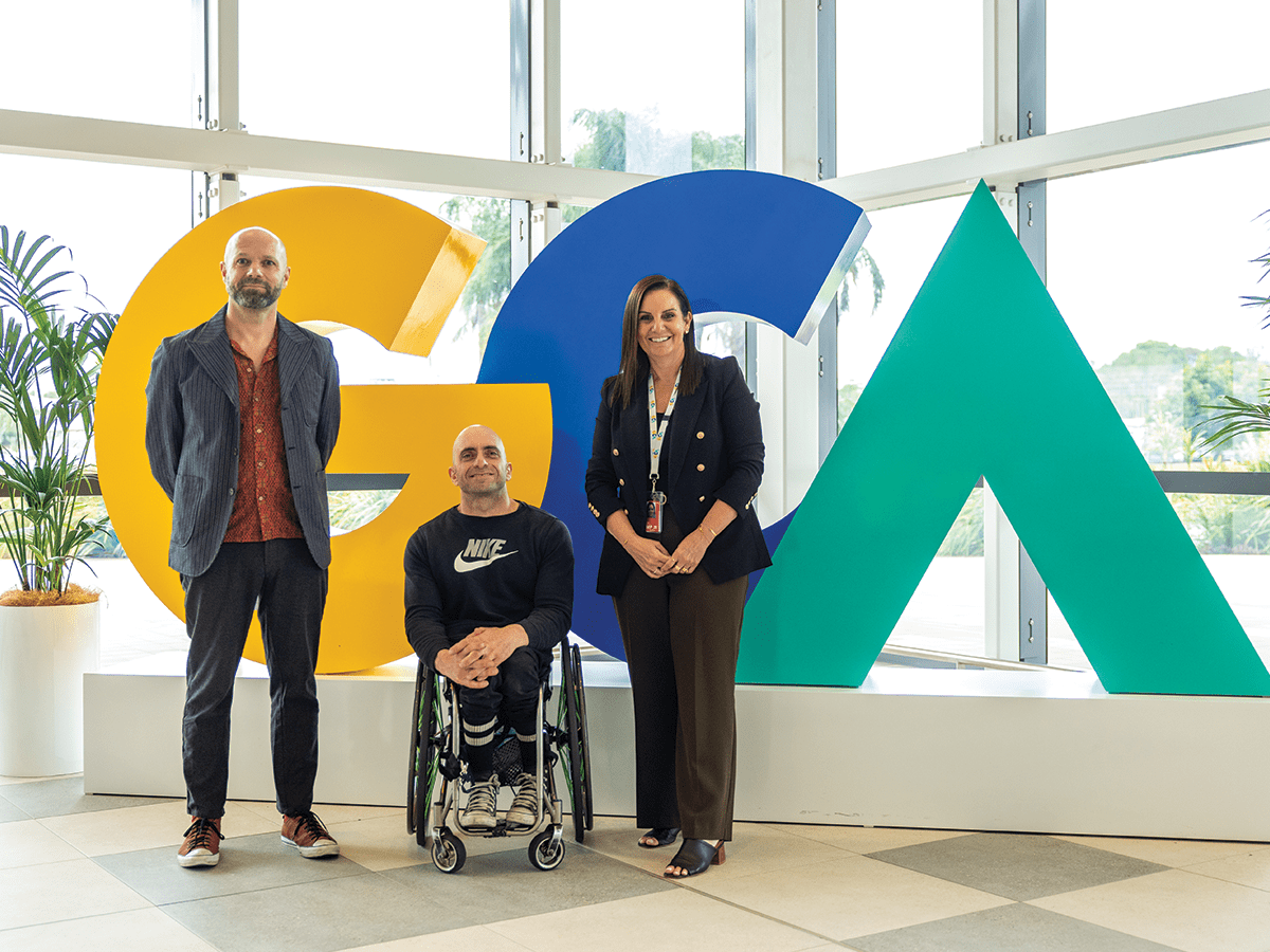 Three people stand in front of a sign of the letters GCA in yellow, blue and green. They are dressed professionally. The middle person is a man in a wheelchair. On his right is a tall man and on his left is a lady.