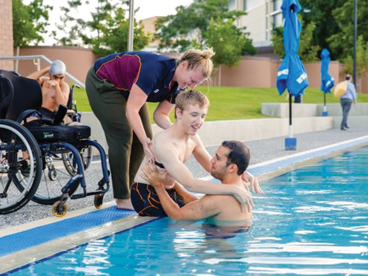 A wheelchair sits at the edge of a swimming pool. A young man is being assisted into the pool by a woman in a maroon shirt, and a shirtless man in the water.