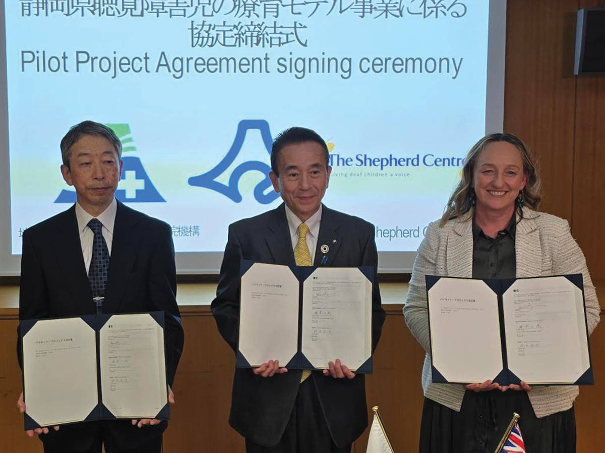 Three people stand in a row, holding official looking papers. There is a presentation on a screen behind them that tells us this is the signing ceremony for the Shepherd Centre's activities in Japan.