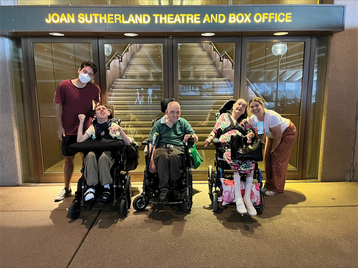 Five people are outside a theatre box office. Three are in wheelchairs. Two are standing next to them. They all look happy.