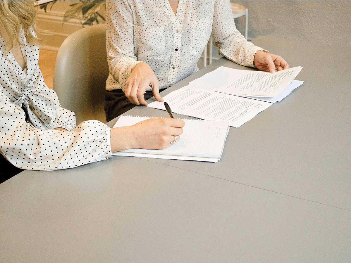 Two ladies sit at a desk. We can only see their hands. They have paper in front of them on a grey table and they seem to be writing on it. They are dressed in business shirts.