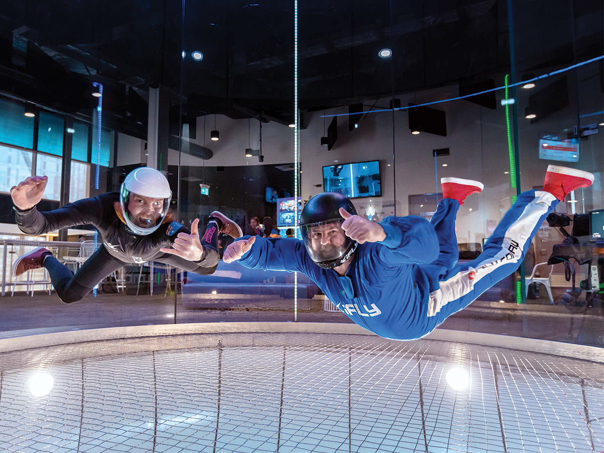 Two people doing indoor skydiving hover in the centre of the photo. They are in jumpsuits and smiling at the camera.