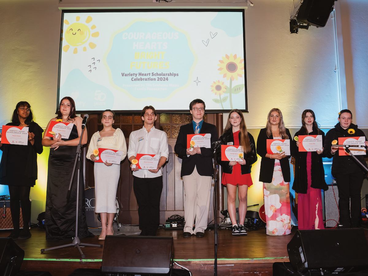 The 2024 Variety scholarship recipients: a group of young boys and girls standing on a stage in front of a projector screen. They are all holding certificates.