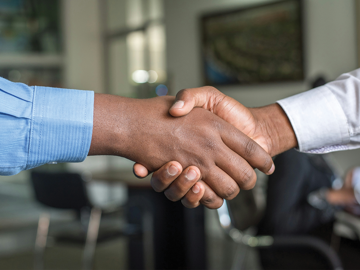 two men with dark skin in business shirts shake hands. We can only see their hands and wrists. They seem to be in an office setting.