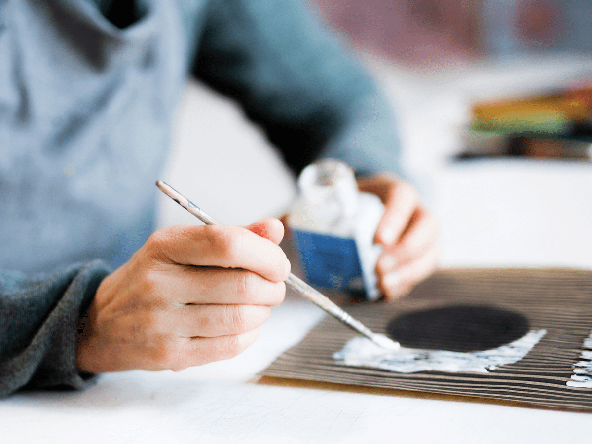 A person holding a paintbrush and bottle of paint, painting an object lying on a table in front of them.