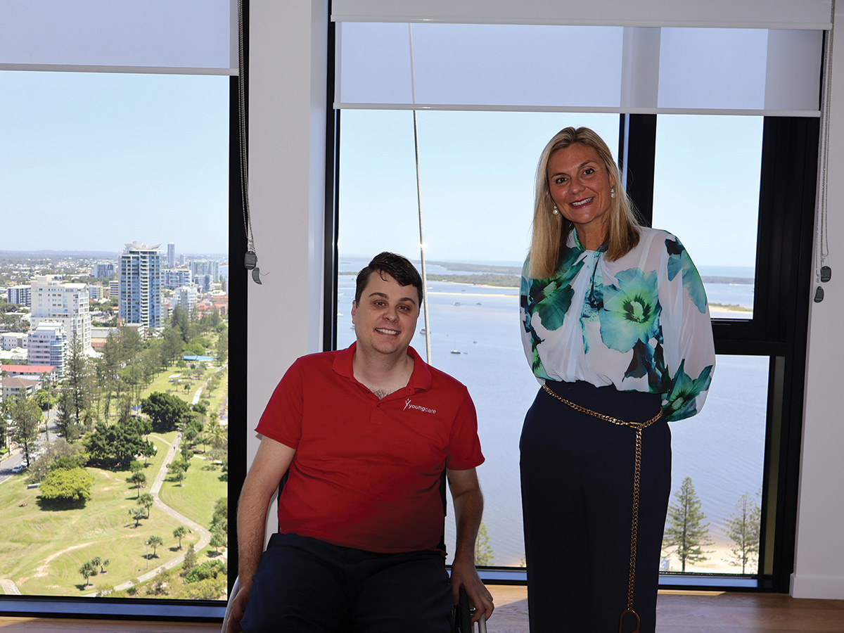 A young man in a wheelchair in a red shirt, and a lady in a blue toned shirt and pants, stand in front of a window with a view out over a city and park.