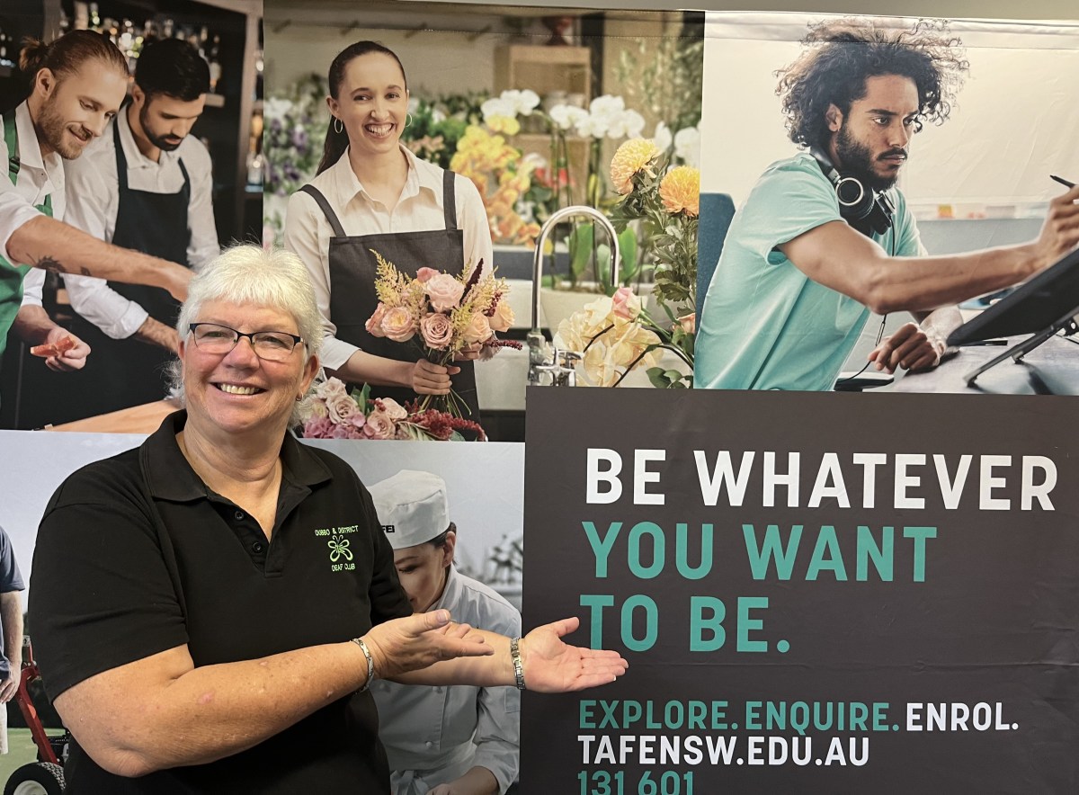 Wendy Daley, an older woman with white hair in a black shirt, gestures towards a sign behind her that says 'be whatever you want to be'. She is smiling brightly.