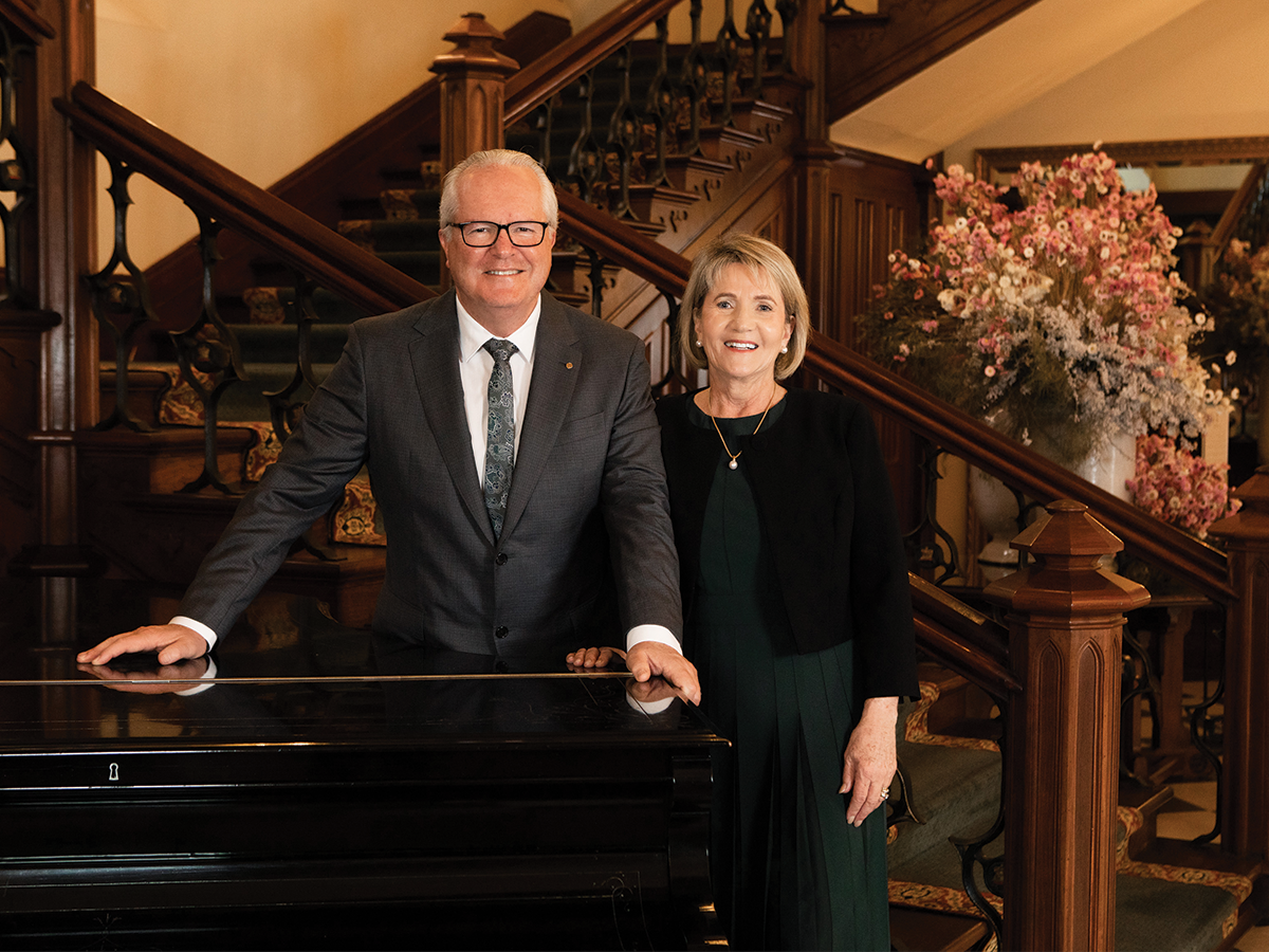 An elderly man and woman stand next to each other in front of a wooden staircase. They are both dressed in dark colours. The man has black glasses on and the lady has blonde hair. They are smiling.