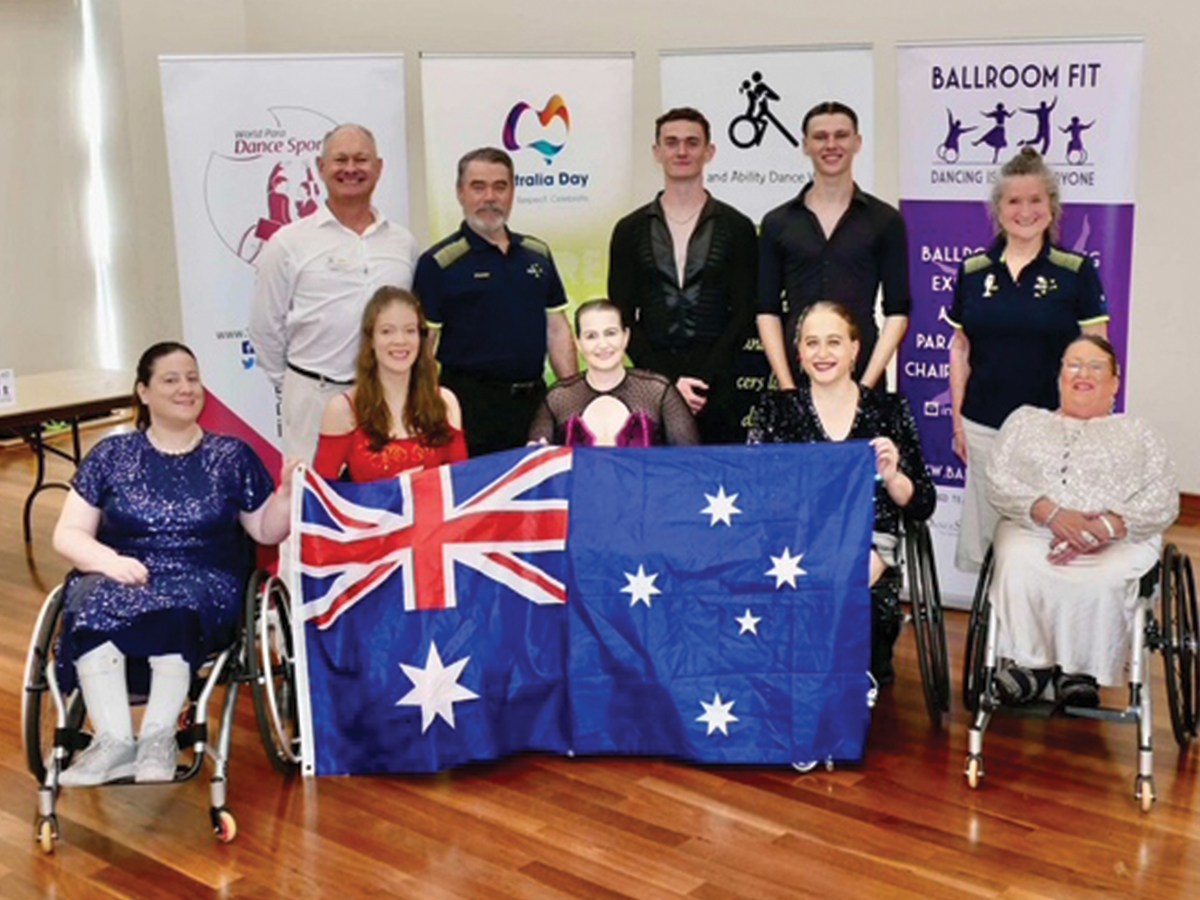 A team of Paradancers, men and women, some in wheelchairs, behind an Australian flag and in front of banners about the para dancers competition.