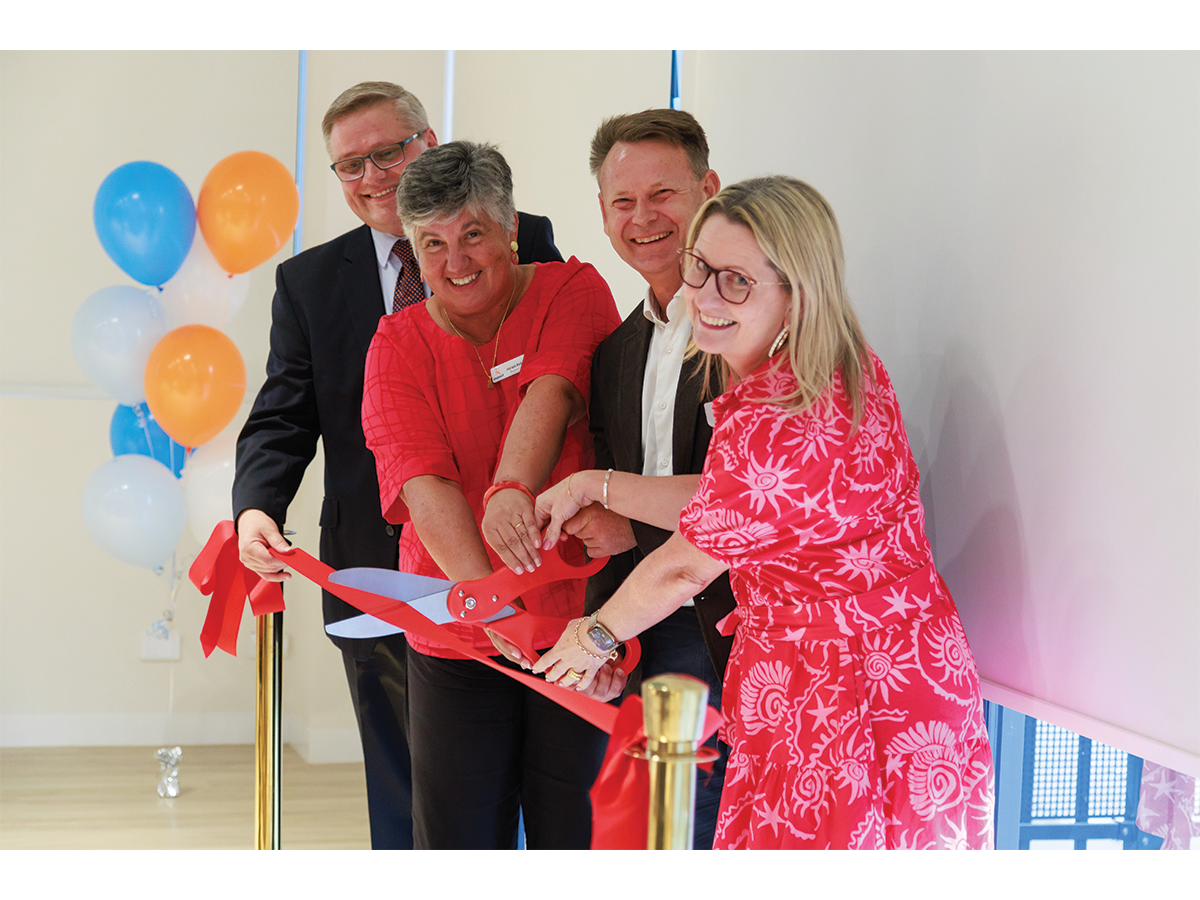 A group of four people wearing bright, professional clothes cut a ribbon together with balloons behind them. They are smiling.